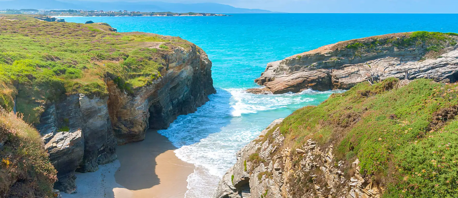 A secluded sandy cove between green cliffs and bright turquoise water on the Galician coast