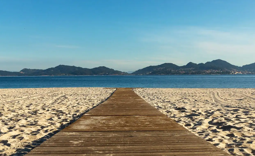 A wooden walkway crossing the pale sand of Praia do Vao towards the calm blue ría